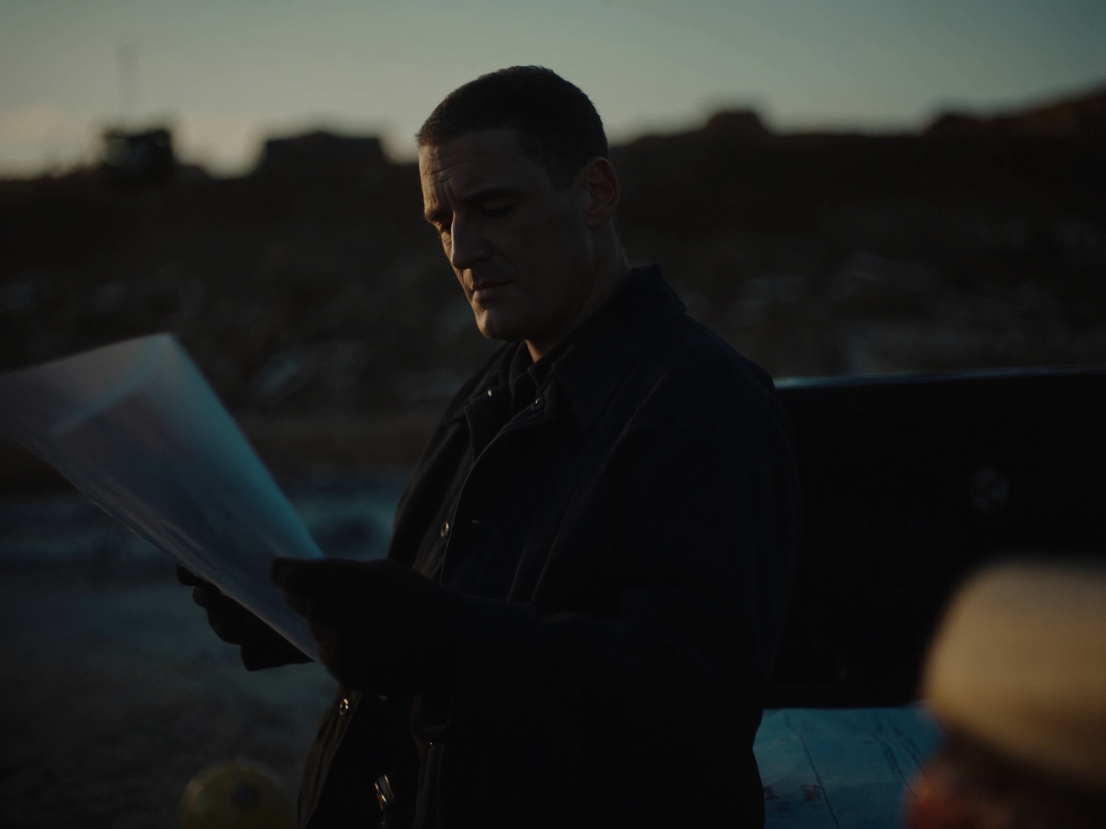 A contractor leans against his truck at dusk, calmly reviewing a set of plans.