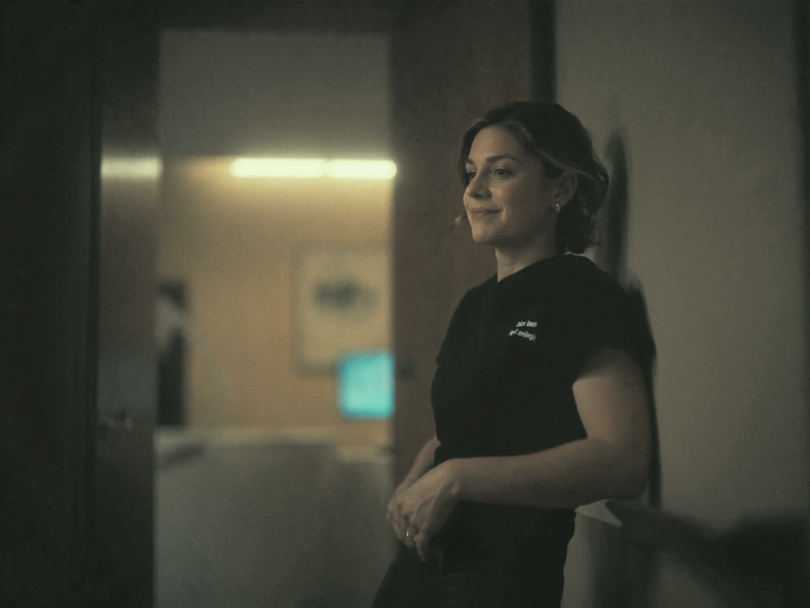 A dentist in scrubs leans against a treatment room doorway with a calm smile.