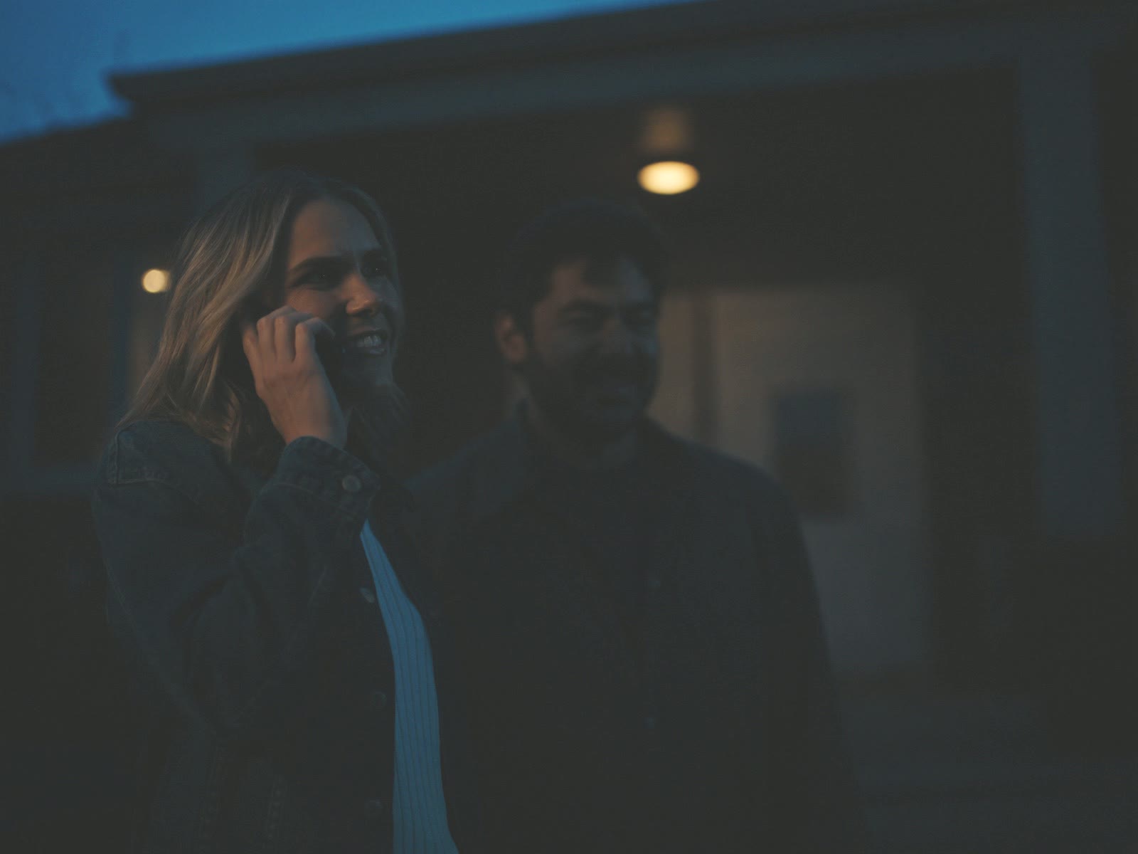 A couple stands in front of a house at dusk, one of them smiling on the phone as their showing is confirmed.