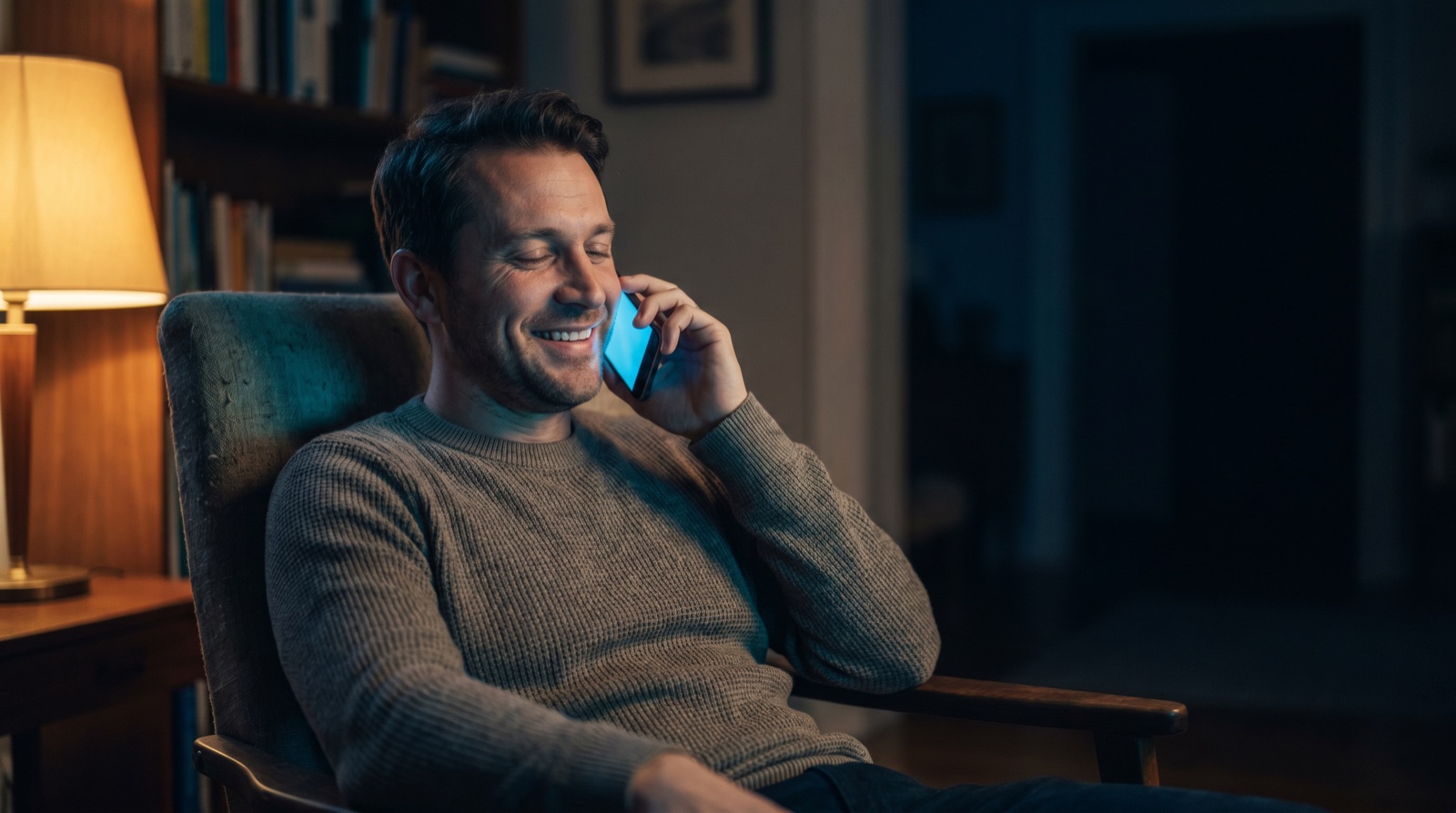 A man at home smiles with relief as he talks on the phone in the evening, warm lamplight beside him.