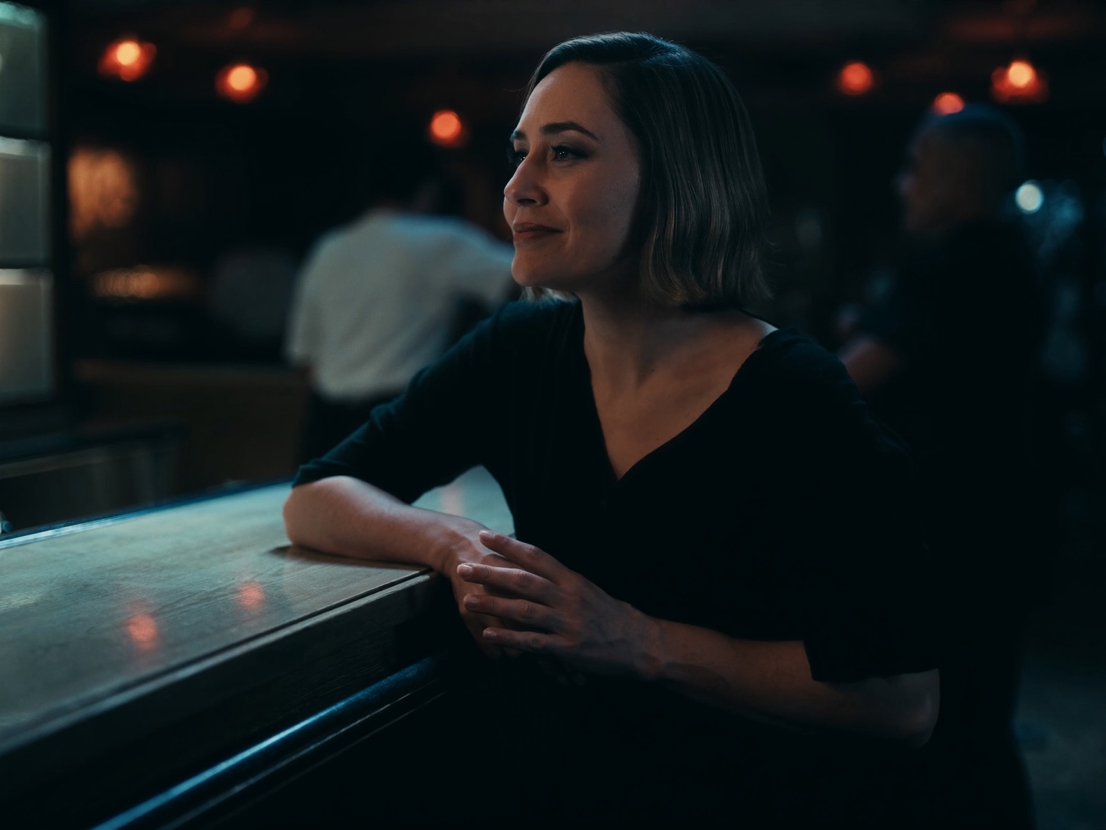 A restaurant owner leans at the bar with a calm smile while evening service runs behind her.