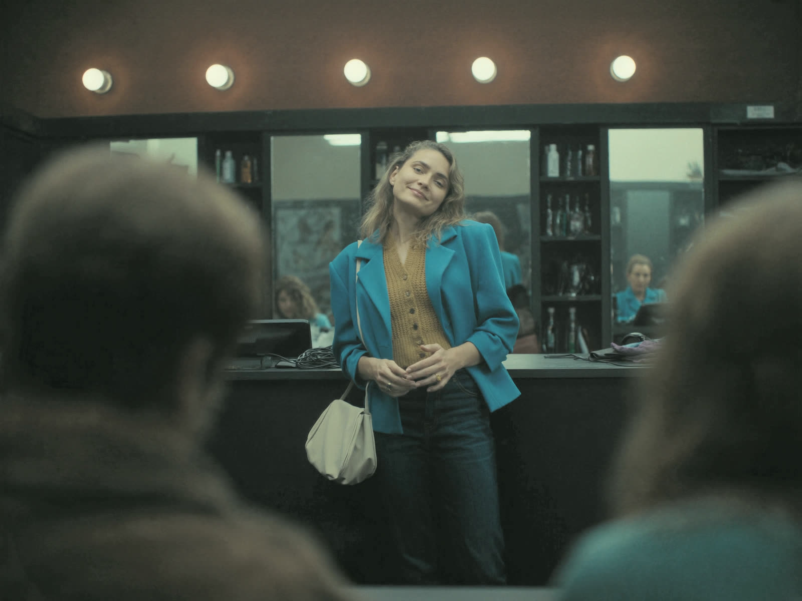 A salon owner stands relaxed at her reception counter between clients, styling stations glowing behind her.