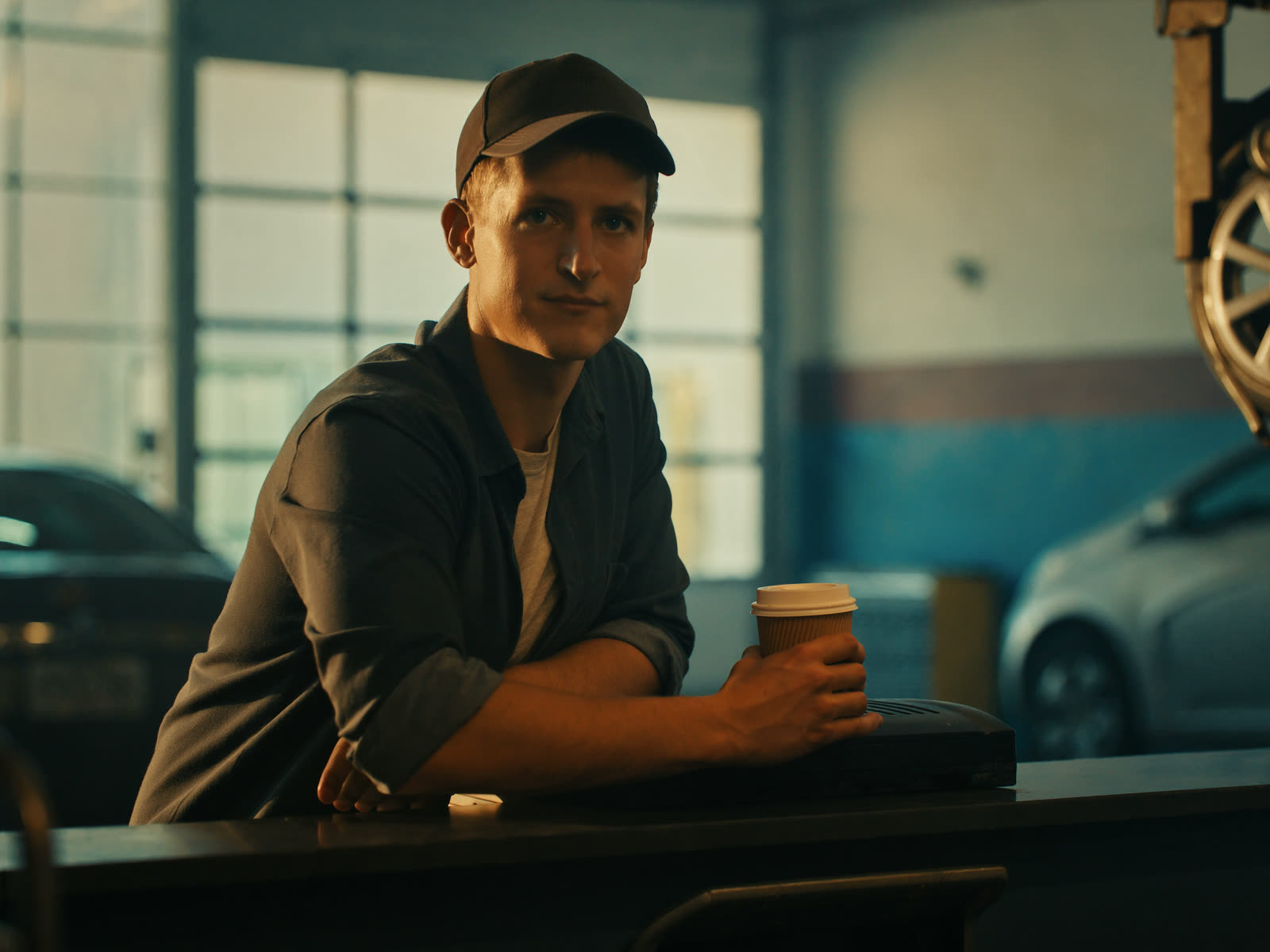 A shop owner leans on his service counter with a morning coffee, calm, the garage bay behind him.